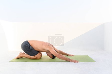 Foto de Hombre 50 practicando yoga, estirando pose, pose infantil, en ropa deportiva en día soleado en la terraza. Foto de alta calidad - Imagen libre de derechos