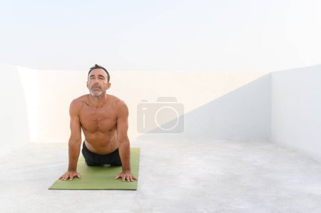 Photo for Mid adult man practicing Upward Facing Dog Pose. Handsome fit male is doing yoga on exercise mat. He is in sportswear with bright background - Royalty Free Image