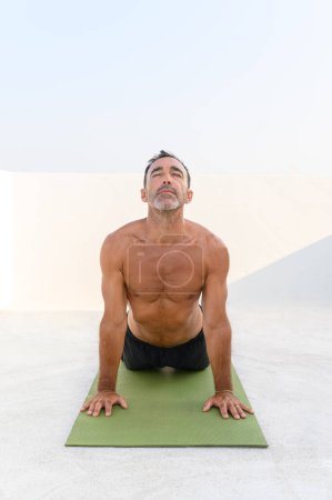 Foto de Hombre adulto practicando Pose boca arriba. Hombre guapo en forma está haciendo yoga en la esterilla de ejercicio. Él está en ropa deportiva con fondo brillante, tiro vertical - Imagen libre de derechos