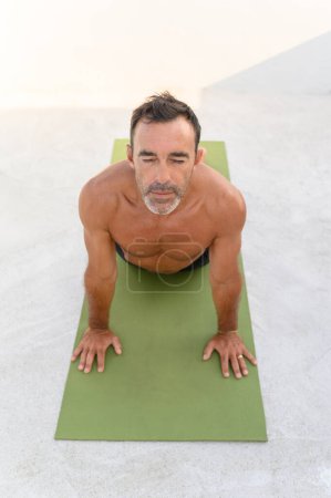 Foto de Hombre adulto practicando Pose boca arriba. Hombre guapo en forma está haciendo yoga en la esterilla de ejercicio. Él está en ropa deportiva con un fondo brillante - Imagen libre de derechos