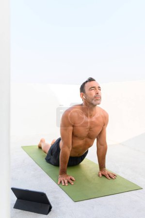 Foto de Hombre adulto practicando Pose boca arriba. Hombre guapo en forma está haciendo yoga en la esterilla de ejercicio junto a su computadora portátil. Él está en ropa deportiva con un fondo brillante - Imagen libre de derechos