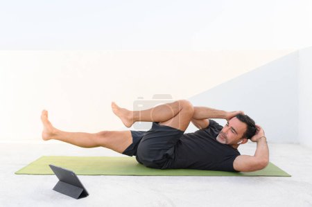 Téléchargez les photos : Homme en bonne santé exercice abdominaux sur le sol. Sporty homme d'âge moyen formation à la maison et regarder tutoriel en ligne sur ordinateur portable, portant noir court et t-shirt photo de haute qualité - en image libre de droit