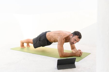 Photo for Side view of sporty strong man 50s doing plank challenge online, wearing black shorts and t-shirt on terrace on a sunny day. yoga mat, earbuds and laptop. High quality photo - Royalty Free Image