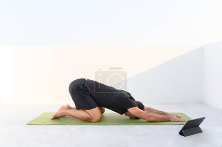 Téléchargez les photos : Homme 50 pratiquer le yoga, pose d'étirement, pose d'enfant, dans des vêtements sportifs par jour ensoleillé sur la terrasse. Photo de haute qualité - en image libre de droit