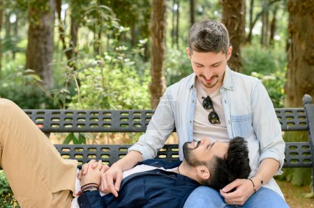 Photo for Young homosexual male chilling on bench on knees of soulmate while looking at each other happily in park in summer - Royalty Free Image