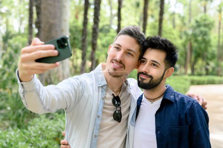 Photo for Content young male and boyfriend taking self portrait on phone while standing in summer green forest and embracing each other - Royalty Free Image