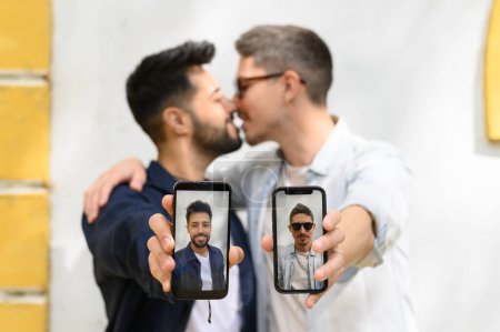 Photo for Gay couple kissing near building wall and outstretching arms with cellphones while demonstrating self portraits on screens in selective focus - Royalty Free Image