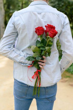 Photo for Back view of unrecognizable male in blue shirt and jeans standing with bouquet of red roses behind back on street - Royalty Free Image