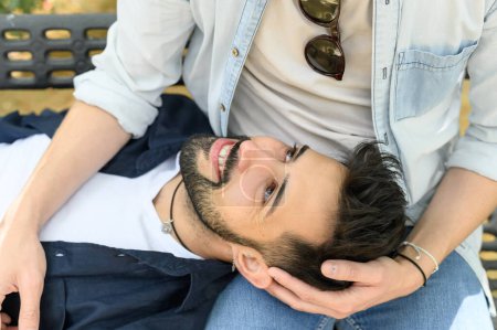 Photo for High angle of cheerful man lying on bench in park and looking up at unrecognizable boyfriend while spending time together - Royalty Free Image