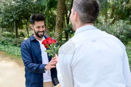 Foto de Positivo barbudo gay tomando ramo de rosas frescas de anónimo novio mientras de pie juntos en verde parque en verano - Imagen libre de derechos