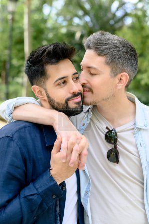 Enamored young guy in casual clothes embracing and kissing bearded Hispanic boyfriend while holding hands against green trees in park