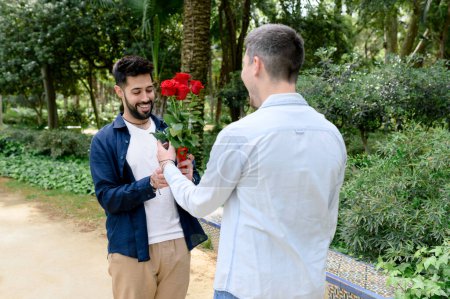 Foto de Hombre irreconocible en traje casual de pie cerca de árboles verdes y dando rosas rojas a novio alegre mientras citas en el jardín de verano - Imagen libre de derechos