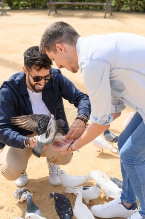 Photo for Positive young male friends in casual outfits smiling and playing with pigeons while feeding birds on sunny sandy ground in park together - Royalty Free Image