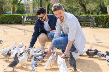 Téléchargez les photos : Heureux jeunes amis masculins en tenues décontractées souriant et nourrissant des pigeons mignons tout en passant du temps ensemble sur un sol sablonneux ensoleillé dans le parc - en image libre de droit