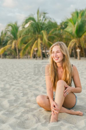Téléchargez les photos : Une jeune femme heureuse aux cheveux longs portant un bikini et un jean court assis et relaxant sur une plage au coucher du soleil dans les arbres tropicaux de fond - en image libre de droit