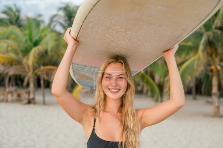 Téléchargez les photos : Joyeux joyeux jeune femme avec de longs cheveux blonds en bikini noir portant planche de surf sur sa tête au coucher du soleil et regardant la caméra - en image libre de droit