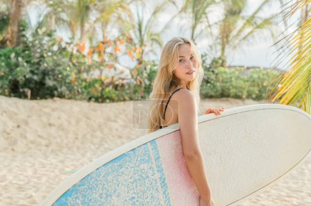 Téléchargez les photos : Jeune fille surfeuse aux cheveux longs blonds portant planche de surf sous le bras tout en marchant sur la plage au coucher du soleil arrière-plan palmiers - en image libre de droit