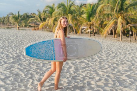 Photo for Young modern woman holding surfboard walking barefoot on sand at beach and looking at camera, background palm trees and sky - Royalty Free Image