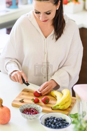 Téléchargez les photos : Concentré jeune femme coupant fraises juteuses fraîches et fruits sur planche à découper en bois tout en cuisinant le petit déjeuner dans la cuisine moderne - en image libre de droit