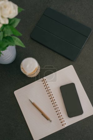 Foto de Vista superior del cuaderno espiral abierto con lápiz y teléfono inteligente con pantalla negra colocada en la mesa cerca del vaso de capuchino espumoso y tableta en la oficina - Imagen libre de derechos