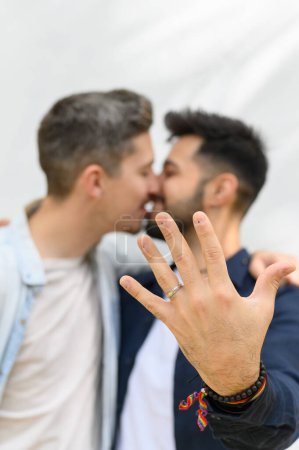 Foto de Vista lateral de hombre gay hispano con cabello oscuro y barba sonriendo y mostrando anillo mientras se besa con novio después de la propuesta - Imagen libre de derechos