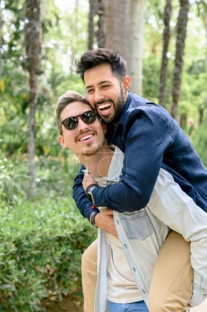 Photo for Side view of delighted beloved ethnic homosexual man in casual clothes and sunglasses giving piggyback ride to laughing boyfriend in lush green park - Royalty Free Image