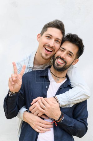 Photo for Smiling bearded boyfriends with short black hair standing near white wall and embracing while showing peace gesture and looking at camera - Royalty Free Image