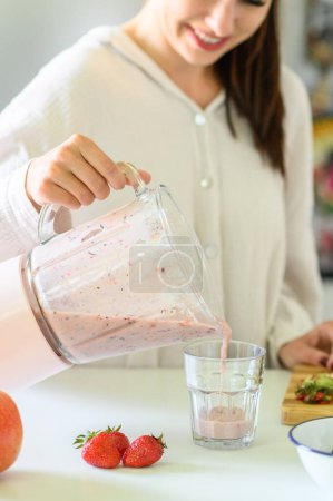 Téléchargez les photos : Crop positive jeune femme en chemise blanche décontractée verser smoothie fraise fraîche dans un verre transparent et souriant tout en préparant un petit déjeuner sain dans la cuisine - en image libre de droit