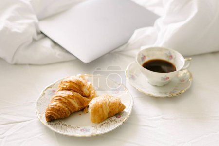 Photo for From above cup of hot aromatic black coffee placed on crumpled bed with laptop and plate of delicious croissant - Royalty Free Image