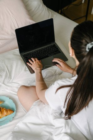 Photo for From above of crop anonymous female sitting on bed with plate of croissant and browsing netbook while working at home - Royalty Free Image