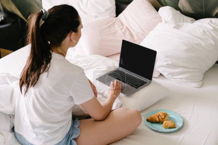 Photo for High angle back view of young female in casual clothes sitting on bed with pillows and croissant snacks while browsing laptop with blank screen - Royalty Free Image