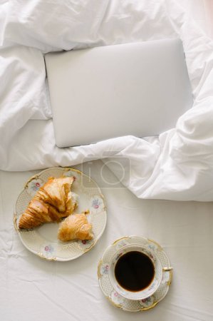 Foto de Vista superior de la taza de café negro colocado en la cama cerca del plato de delicioso croissant con computadora portátil en la sábana arrugada sobre la cama en el dormitorio - Imagen libre de derechos