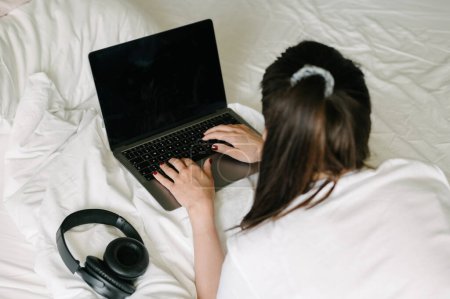 Foto de Vista trasera de gran angular de una freelancer anónima acostada en la cama con auriculares y escribiendo en el portátil mientras trabaja en un proyecto remoto en casa - Imagen libre de derechos