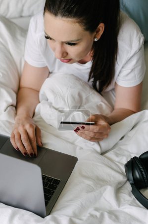 Photo for High angle of young female in casual clothes lying on bed and browsing laptop while holding credit card at home - Royalty Free Image