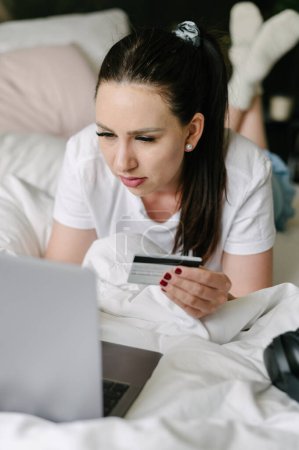 Foto de Mujer joven enfocada en ropa casual con cabello oscuro usando computadora portátil mientras sostiene la tarjeta de crédito en la cama en casa - Imagen libre de derechos
