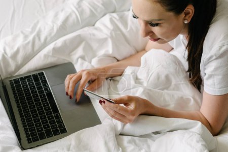 Photo for High angle of crop young female in casual clothes lying on bed and shopping on laptop with bank card while resting in bedroom - Royalty Free Image