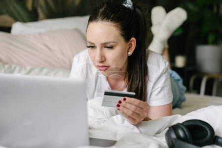 Foto de Mujer concentrada en ropa casual navegador portátil mientras sostiene la tarjeta de crédito tumbado en la cama en casa - Imagen libre de derechos