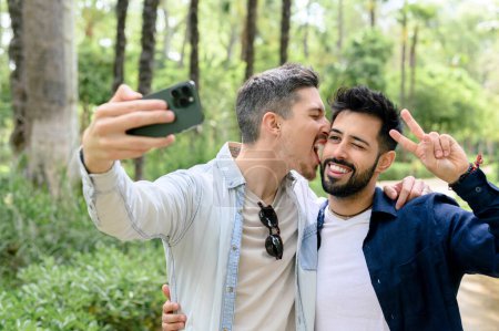 Foto de Feliz joven barbudo y su novio en ropa casual sonriendo y tomando selfie mientras está de pie en el bosque verde en el día soleado - Imagen libre de derechos