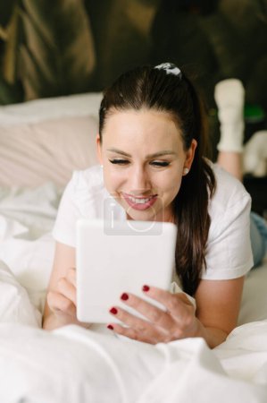 Foto de Joven mujer alegre con el pelo oscuro acostado en la cama y tableta de navegación mientras descansa en casa durante el día de fin de semana - Imagen libre de derechos