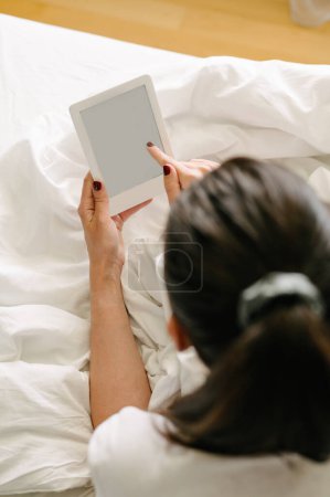 Téléchargez les photos : Vue de dessus vers l'arrière de la femelle anonyme couchée sur le lit et utilisant une tablette avec écran blanc à la maison - en image libre de droit
