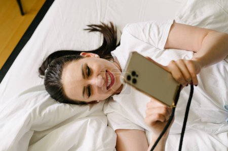 Téléchargez les photos : D'en haut de joyeuse jeune femme couchée sur le lit et prenant selfie sur smartphone dans la chambre à coucher à la maison pendant le week-end - en image libre de droit