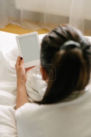 Téléchargez les photos : Vue de dessus vers l'arrière de la femelle anonyme couchée sur le lit et le livre de lecture sur lecteur électronique avec écran vide à la maison - en image libre de droit