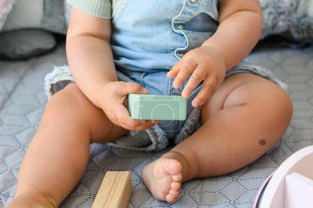 Photo for Crop anonymous baby in casual clothes sitting on bed holding toy block while playing in bedroom at home - Royalty Free Image