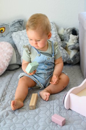 Photo for Full body of adorable baby in casual clothes sitting on soft bed playing with wooden toy blocks in bedroom at home - Royalty Free Image