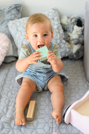 Téléchargez les photos : Corps complet de bébé adorable en vêtements décontractés mordant bloc jouet en bois tout en étant assis sur le lit à la maison en regardant la caméra - en image libre de droit