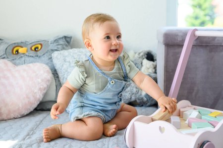 Photo for Full length of cute little baby in casual clothes playing with toy blocks in cart while sitting on soft bed at home - Royalty Free Image