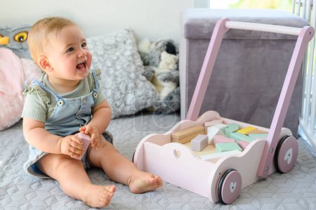 Photo for Adorable baby in casual clothes sitting on bed and playing with toy blocks near cart while spending time at home and looking away - Royalty Free Image