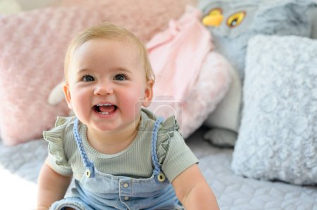 Photo for Portrait of adorable little baby in casual clothes smiling and looking at camera while sitting on bed in bedroom at home - Royalty Free Image