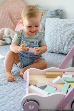 Téléchargez les photos : Joyeux adorable bébé fille jouer avec des blocs de bois près chariot de jouet sur le lit à la maison - en image libre de droit