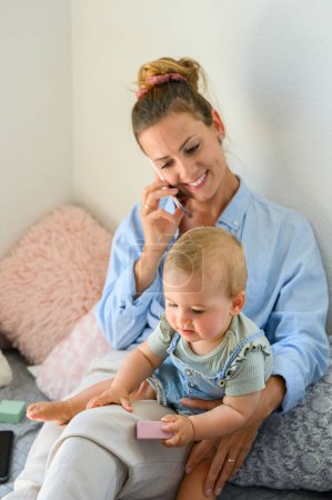 Photo for Delighted young mother in casual wear smiling and having phone conversation on bed with cute toddler daughter while spending time together during weekend - Royalty Free Image